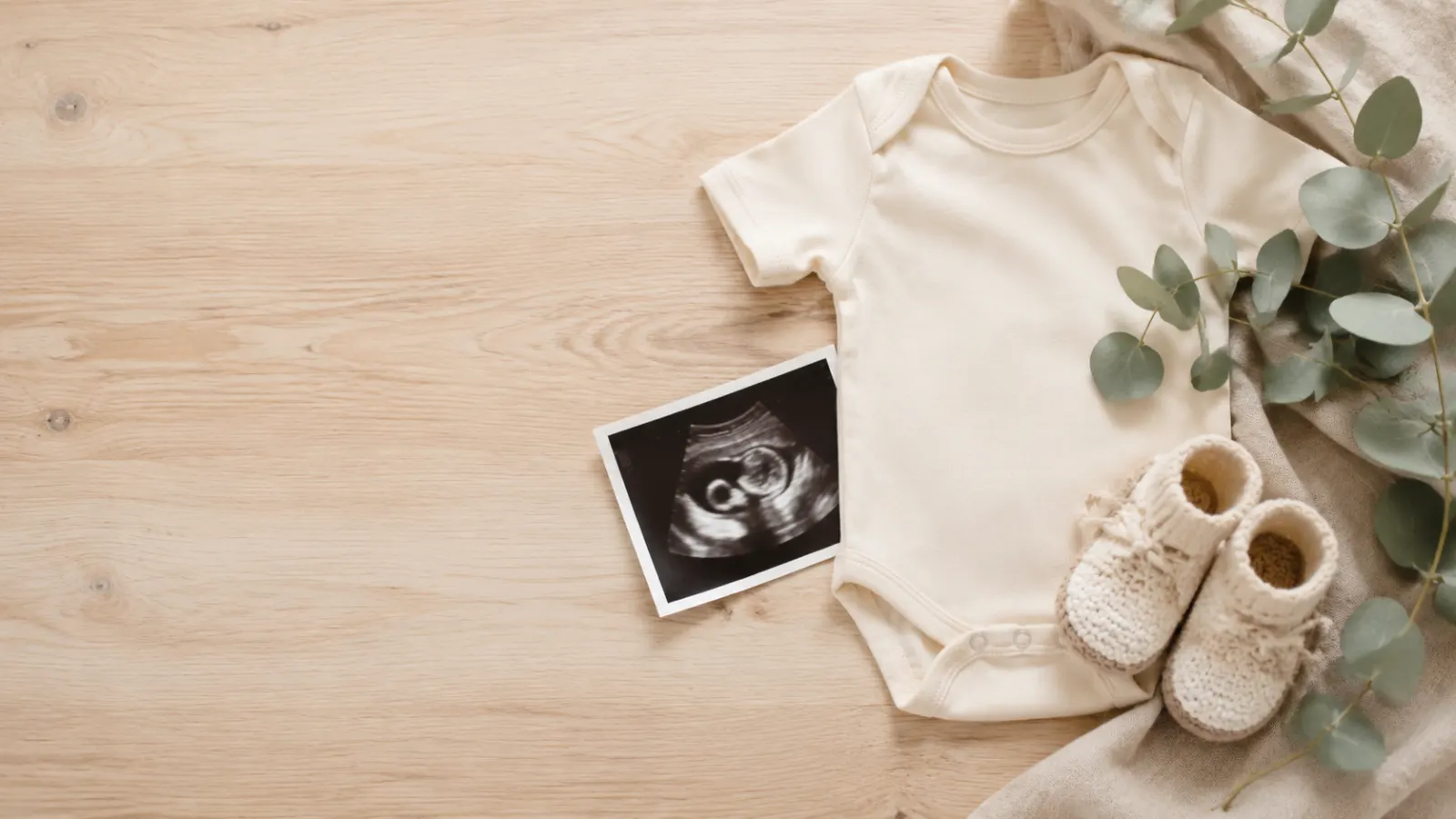 Neutral baby onesie, knitted booties, ultrasound photo, and eucalyptus arranged for a baby announcement.