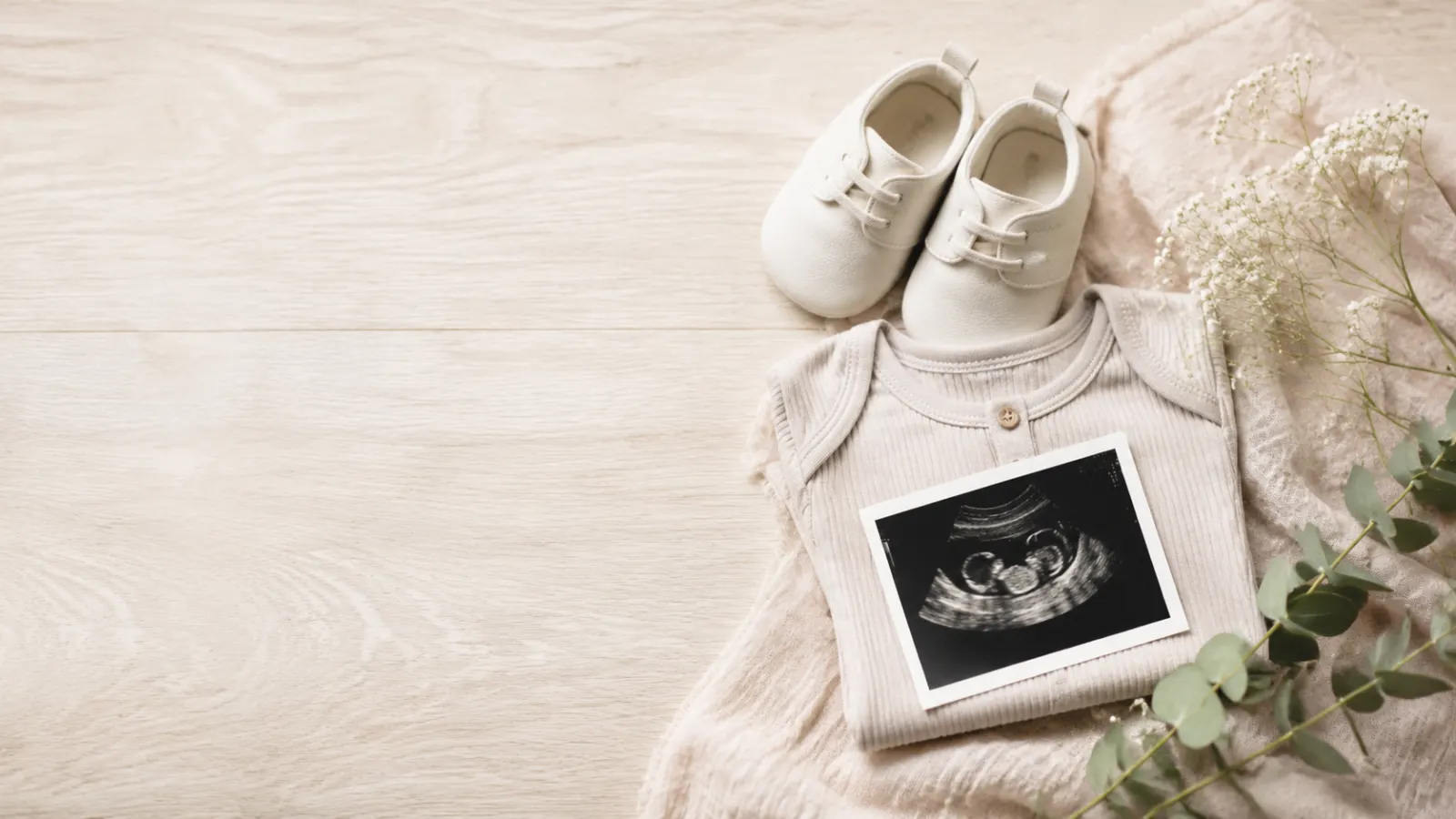 Neutral pregnancy announcement with baby shoes, a folded onesie, flowers, and an ultrasound photo.