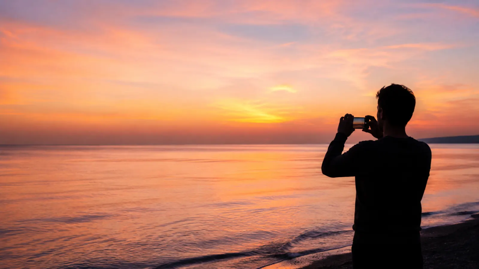 Person photographing a colorful sunset over a calm ocean.