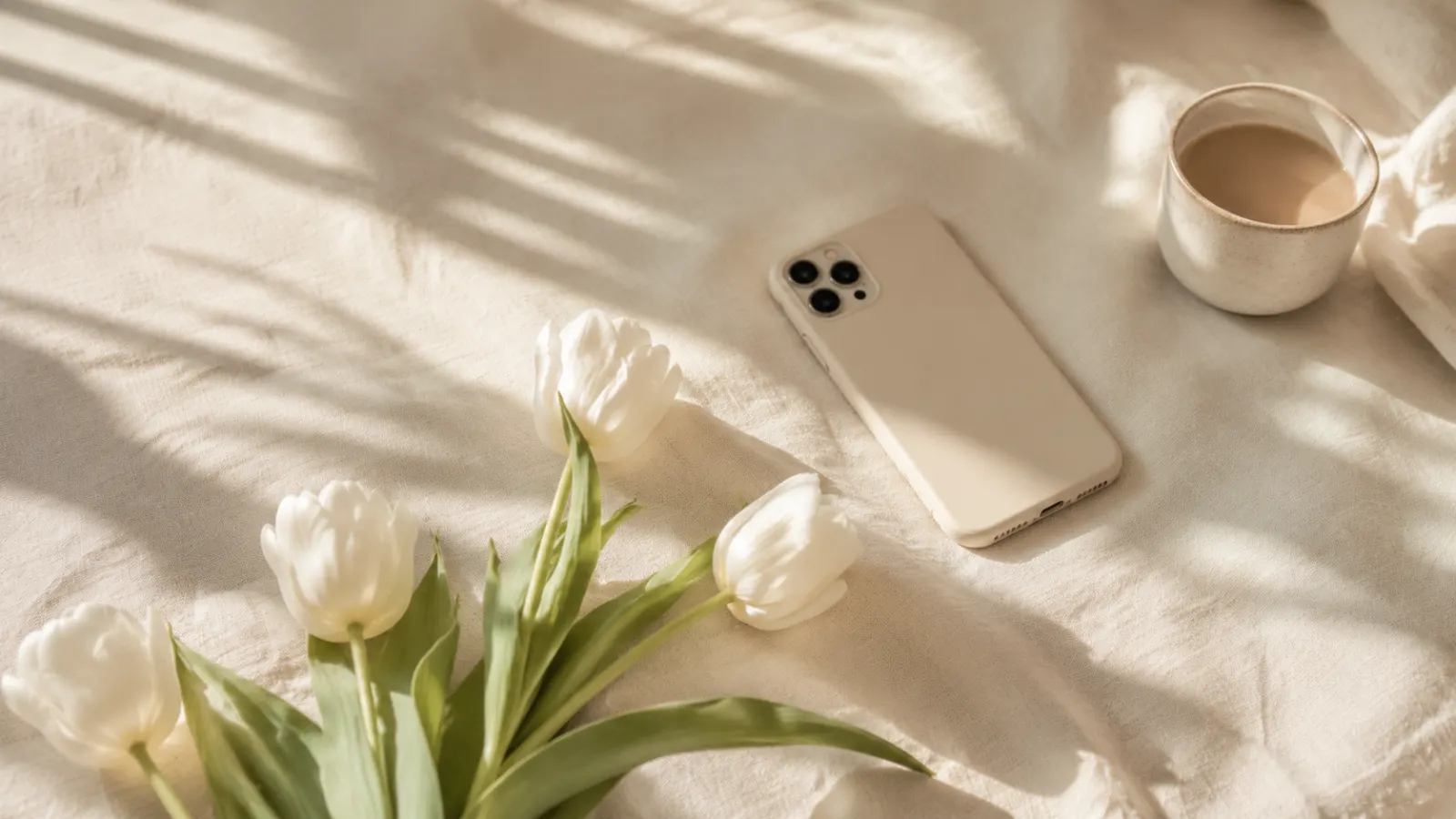 White tulips, coffee, and a smartphone arranged on cream linen in warm morning light.